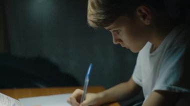 Genç bir çocuk akşamları masa başında ders çalışıyor. Çocuk deftere yazı yazıyor. Genç erkek çocuk odasında masasında ev ödevini yapmaya çok konsantre. Çocuklar ve eğitim. Yüksek kalite 4k görüntü