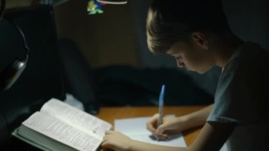 Genç bir çocuk akşamları masa başında ders çalışıyor. Çocuk deftere yazı yazıyor. Genç erkek çocuk odasında masasında ev ödevini yapmaya çok konsantre. Çocuklar ve eğitim. Yüksek kalite 4k görüntü