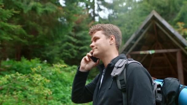 一位英俊的男性游客 一个人在森林里 在森林房子的背景下 用智能手机说话 旅行和旅游概念 — 图库视频影像