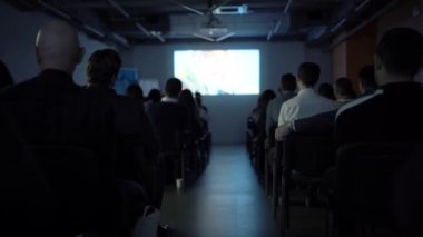 Karanlıkta geniş bir seyirci kitlesi önünde ekranda bir sunum gösteriliyor. İzleyicinin arkasından görüntüle. Eğitim, konferans, forum. Topluluk önünde konuşma konsepti.