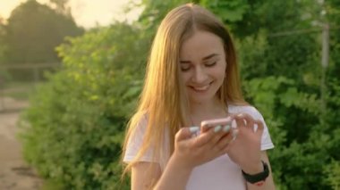Parkta cep telefonunda yazan güzel bir kadının portresi. Kız elinde bir cep telefonu tutuyor, gülümsüyor ve gülüyor. Sosyal ağlarda iletişim.