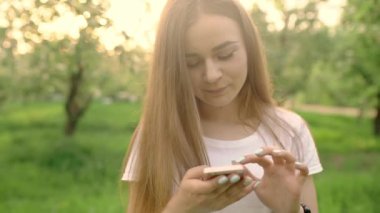 Parkta cep telefonunda yazan güzel bir kadının portresi. Kız elinde bir cep telefonu tutuyor, gülümsüyor ve gülüyor. Sosyal ağlarda iletişim.