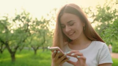 Parkta cep telefonunda yazan güzel bir kadının portresi. Kız elinde bir cep telefonu tutuyor, gülümsüyor ve gülüyor. Sosyal ağlarda iletişim.