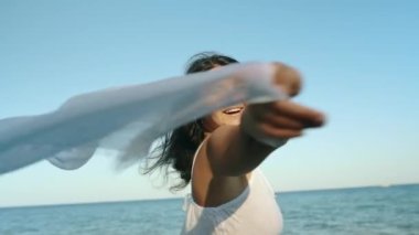A brunette in a white dress holds a fluttering pareo in her hands and enjoys a sunny day. Freedom concept. Tourist on the islands