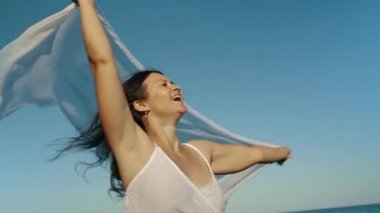 A brunette in a white dress holds a fluttering pareo in her hands and enjoys a sunny day. Freedom concept. Tourist on the islands