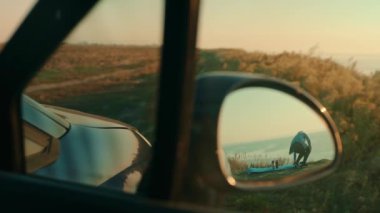 In the side rear-view mirror of the car, a tourist girl is displayed, who sat down to admire the sea sunset. Adventure and travel concept. Travel by car