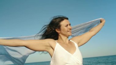 A brunette in a white dress holds a fluttering pareo in her hands and enjoys a sunny day. Freedom concept. Tourist on the islands