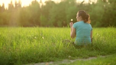 A pretty girl blows on a ripe dandelion while sitting on the grass in the evening against the backdrop of the setting sun. Outdoor recreation. View from the back. 4K slow motion video 
