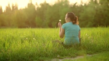 A pretty girl blows on a ripe dandelion while sitting on the grass in the evening against the backdrop of the setting sun. Outdoor recreation. View from the back. 4K slow motion video