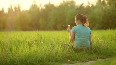A pretty girl blows on a ripe dandelion while sitting on the grass in the evening against the backdrop of the setting sun. Outdoor recreation. View from the back. 4K slow motion video