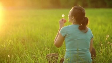 Against the background of the setting sun, a young girl blows on a ripe dandelion, sitting on the grass in the evening. The fluff slowly flies through the air.View from the back. 4K slow motion video