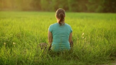 A pretty girl blows on a ripe dandelion while sitting on the grass in the evening against the backdrop of the setting sun. Outdoor recreation. View from the back. 4K slow motion video