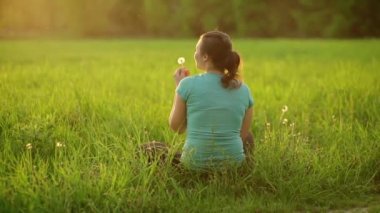 Against the background of the setting sun, a young girl blows on a ripe dandelion, sitting on the grass in the evening. The fluff slowly flies through the air.View from the back. 4K slow motion video