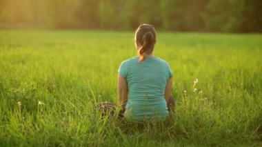 Against the background of the setting sun, a young girl blows on a ripe dandelion, sitting on the grass in the evening. The fluff slowly flies through the air.View from the back. 4K slow motion video