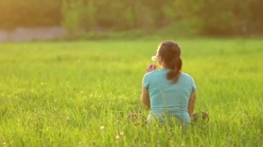 A pretty girl blows on a ripe dandelion while sitting on the grass in the evening against the backdrop of the setting sun. Outdoor recreation. View from the back. 4K slow motion video