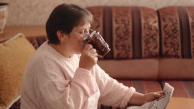 Pretty middle-aged woman in glasses reading a newspaper drinks tea or coffee from a cup while relaxing on a sofa in the living room. Leisure. News source. World pandemic. 4k