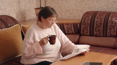 Pretty middle-aged woman in glasses reading a newspaper drinks tea or coffee from a cup while relaxing on a sofa in the living room. Leisure. News source. World pandemic. 4k
