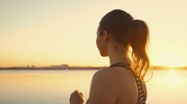 An attractive girl drinks water from a bottle during a training break on the embankment in the sunset. The athlete replenishes the water balance after running.High quality 4k footage
