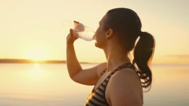 A tired sportswoman replenishes her water balance after a fitness workout in the sun near a pond.Relaxed girl drinks water from a bottle in nature at sunset by the lake.High quality 4k footage