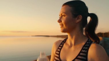 Satisfied with the result and tired, the athlete replenishes the water balance after a fitness workout in the sun by the pond.Relaxed girl drinks water from a bottle in nature at sunset by the lake.4k