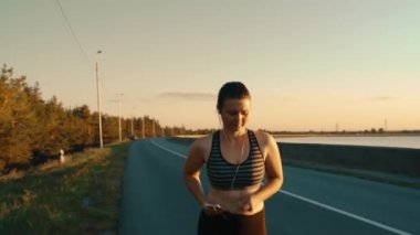 Portrait of a beautiful girl who put on headphones started running along the road at sunset. Healthy lifestyle. High-quality shooting in 4k format.