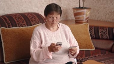 A woman holds a bank card and using the phone, makes purchases at home. Using a credit card online. Online shopping or making payments. Online shopping. High quality 4k footage