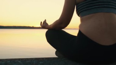 Side view of a young girl practicing yoga and meditation, sitting in a lotus position by the lake in the rays of the setting sun. High quality 4k footage