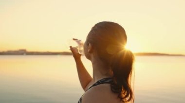 An attractive girl drinks water from a bottle during a training break on the embankment in the sunset. The athlete replenishes the water balance after running. High quality 4k footage