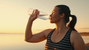 An attractive girl drinks water from a bottle during a training break on the embankment in the sunset. The athlete replenishes the water balance after running.High quality 4k footage