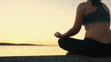 Side view of a young girl practicing yoga and meditation, sitting in a lotus position by the lake in the rays of the setting sun. High quality 4k footage