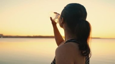 The athlete replenishes the water balance after running on the embankment in the rays of sunset.Portrait of an athlete girl drinking water from a bottle at sunset by the lake.High quality 4k footage