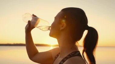 A tired sportswoman replenishes her water balance after a fitness workout in the sun near a pond.Relaxed girl drinks water from a bottle in nature at sunset by the lake.High quality 4k footage