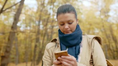 Sonbahar parkında cep telefonuyla duran, e-posta atan, akıllı telefon kullanarak sosyal ağlarda iletişim kuran, internette veya mesajlaşan genç bir kız. Temiz havada yürüyün. Yüksek kalite 4K görüntü.