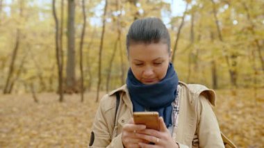 Sonbahar parkında cep telefonuyla duran, e-posta atan, akıllı telefon kullanarak sosyal ağlarda iletişim kuran, internette veya mesajlaşan genç bir kız. Temiz havada yürüyün. Yüksek kalite 4K görüntü.