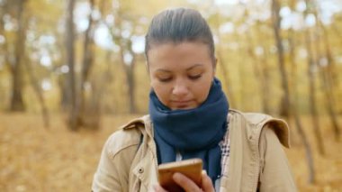 Güz parkında cep telefonuyla duran genç bir kız mesaj yazıyor. İnternetten gelen haberleri telefonla okuyor. Taze sahilde yürüyün. Yüksek kalite 4K görüntü.