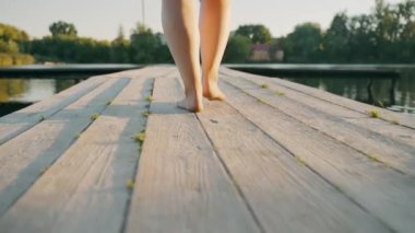 Açık renk elbiseli genç bir kadın göl kenarındaki eski bir ahşap köprüde yalınayak yürüyor. Yaz tatili. Rahat ol. Yüksek kalite 4k görüntü