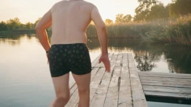 Gençler ahşap bir iskeleden göle atlayıp arkalarında su damlatırlar. Arka manzara. Neşeli bir açık hava eğlencesi. Hile yaparak suya atlamak. Yüksek kalite 4K görüntü.