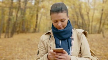 Güz parkında cep telefonuyla duran genç bir kız mesaj yazıyor. İnternetten gelen haberleri telefonla okuyor. Taze sahilde yürüyün. Yüksek kalite 4K görüntü.