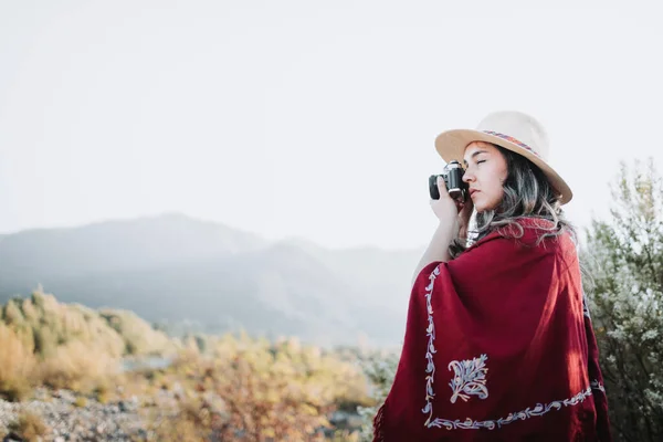 Ambiente de verano mujer joven con un poncho rojo y un sombrero, utilizando una cámara vintage para tomar fotos en un espacio natural. — Foto de Stock