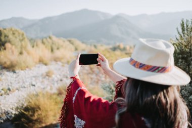 Şapkalı ve kırmızı pançolu bir kadın ve cep telefonuyla doğal ortamlarda fotoğraf çekiyor..
