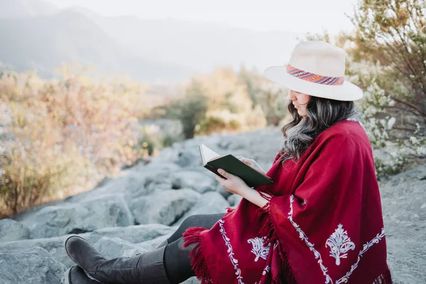 Mujer latina joven usando un sombrero y un poncho rojo, sentada al aire libre y leyendo un libro. Copiar espacio. — Foto de Stock
