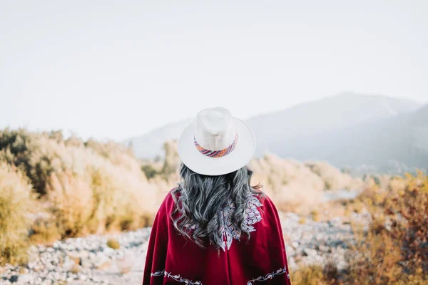 Traditional latin american woman wearing a red poncho and a hat seen from behind, in a natural space. Womens empowerment — Stock Photo, Image