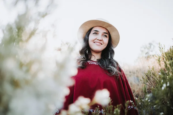 Traditional latin american woman wearing a red poncho and a hat seen from behind, in a natural space. Womens empowerment