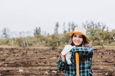 Genç, gülümseyen Latin köylü kadın bahçe eldivenleri kullanıyor ve küreğe yaslanıyor..