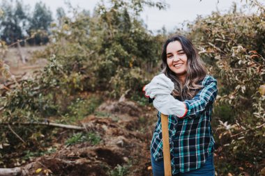 Genç, gülümseyen Latin köylü kadın bahçe eldivenleri kullanıyor ve küreğe yaslanıyor..