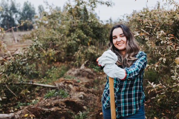 Genç, gülümseyen Latin köylü kadın bahçe eldivenleri kullanıyor ve küreğe yaslanıyor..