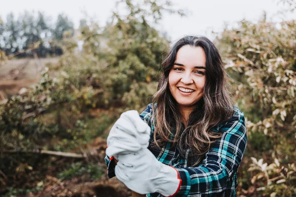 Gülümseyen bir Latin köylü kadının orta boy bir fotoğrafı. Bahçe eldivenleri ve küreklere yaslanmış..
