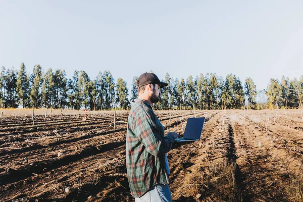 Młody latynoski farmer teleportujący się na swoim laptopie na środku swojej farmy. Zrównoważony rozwój rolnictwa — Zdjęcie stockowe