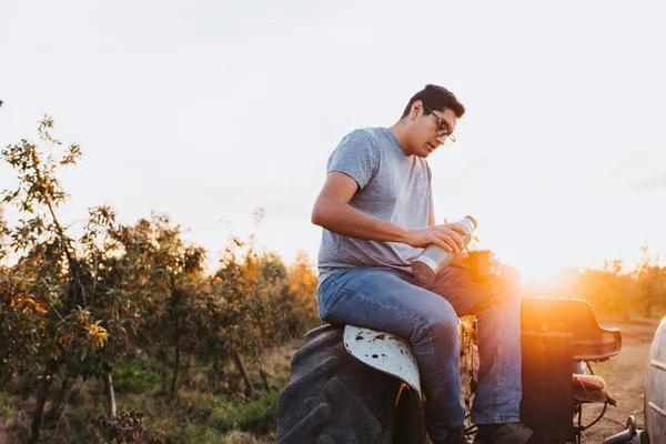 Young latin man drinking mate and relaxing, in a rural space, on top of an old little tractor. Hot beverage — Stock Fotó