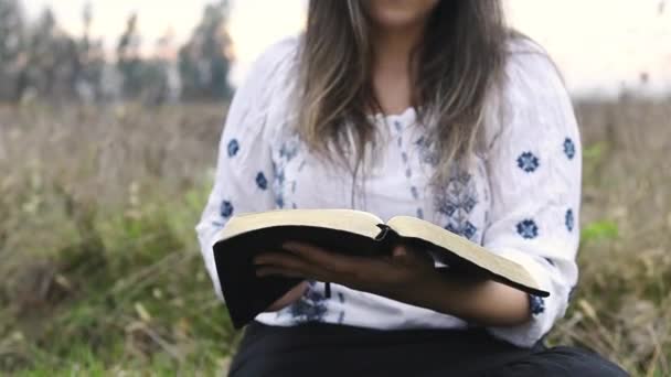 Vrouw met bril zittend buiten op het gras houden en lezen open Bijbel — Stockvideo